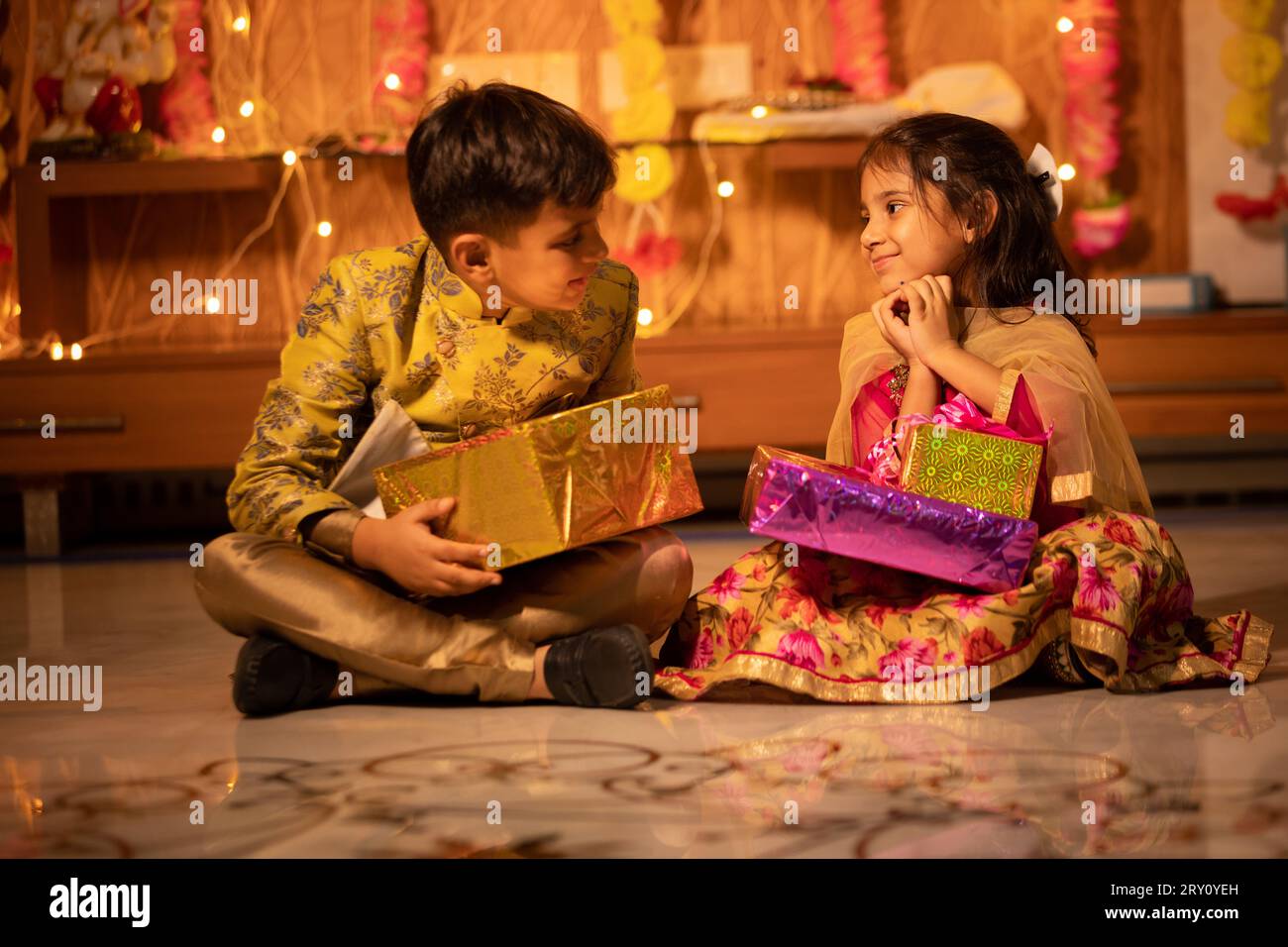 Happy little indian kids brother and sister holding gift boxes in hand celebrating Diwali while sitting on floor, Raksha Bandhan, Bhai Dooj concept. Stock Photo