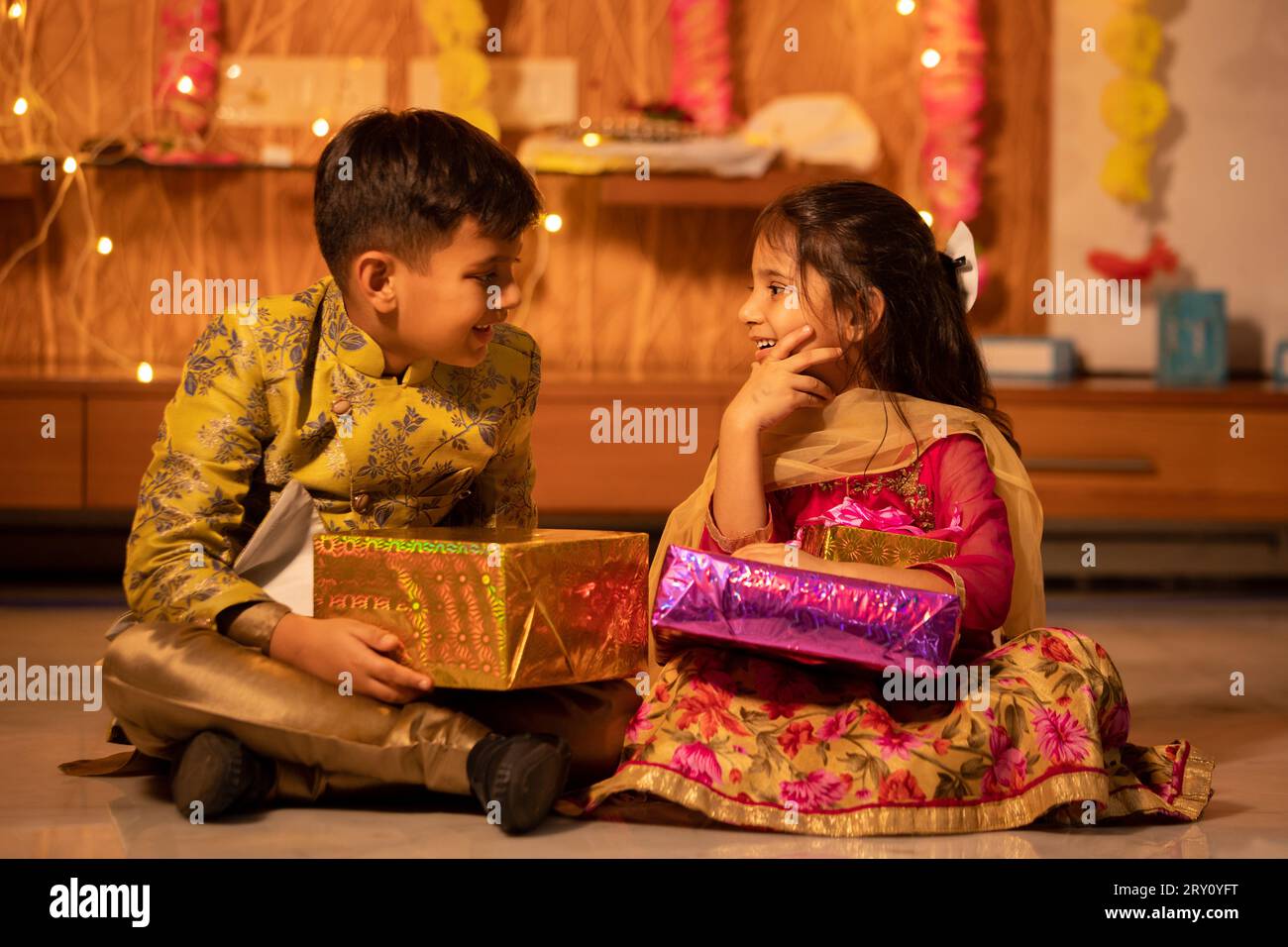 Happy little indian kids brother and sister holding gift boxes in hand celebrating Diwali while sitting on floor, Raksha Bandhan, Bhai Dooj concept. Stock Photo