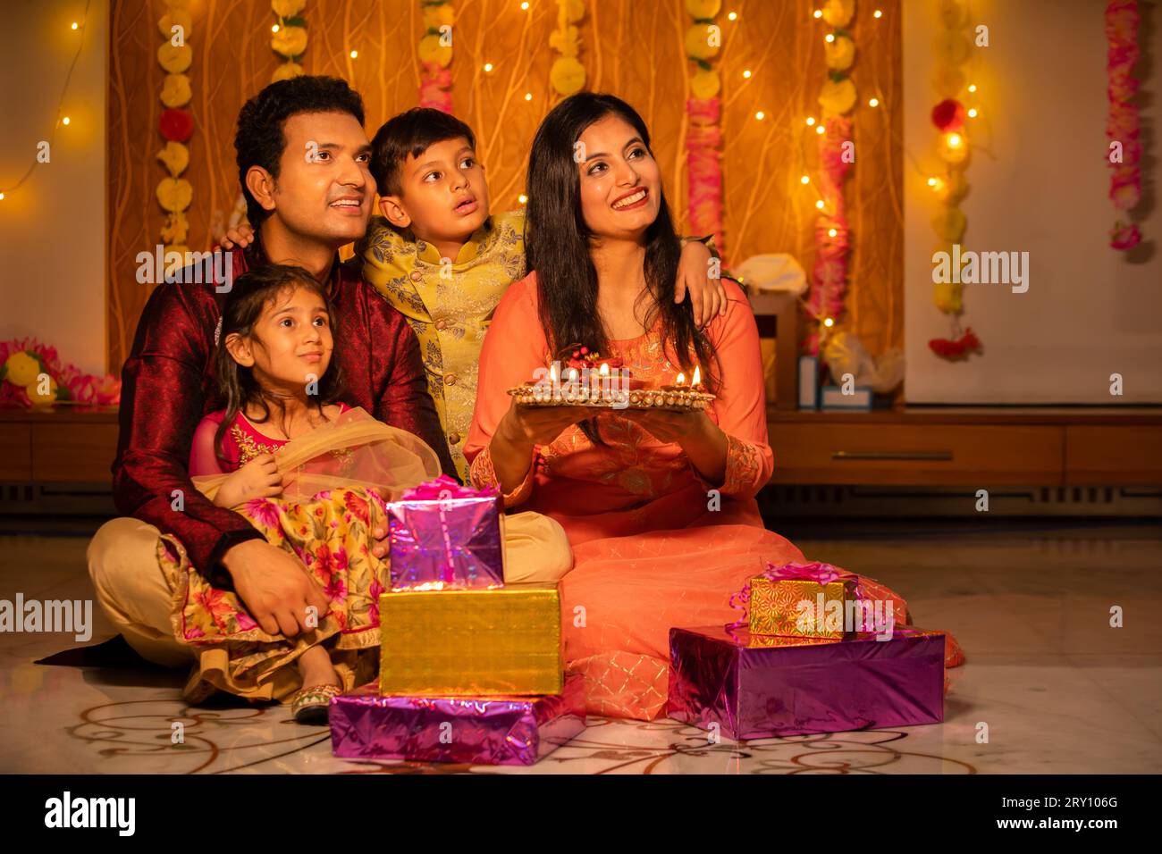 Happy young Indian family in traditional dress with lots of gifts around sitting on floor celebrating diwali festival at home. Stock Photo