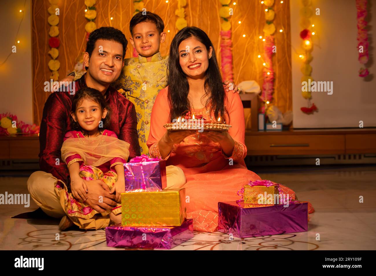 Portrait of happy young Indian family in traditional dress with lots of gifts around sitting on floor celebrating diwali festival at home. Stock Photo