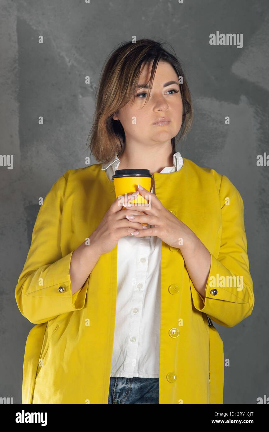 Thoughtful lady with brown short hair standing on gray background and holding yellow disposable coffee cup in hands. Young woman wearing bright xantho Stock Photo