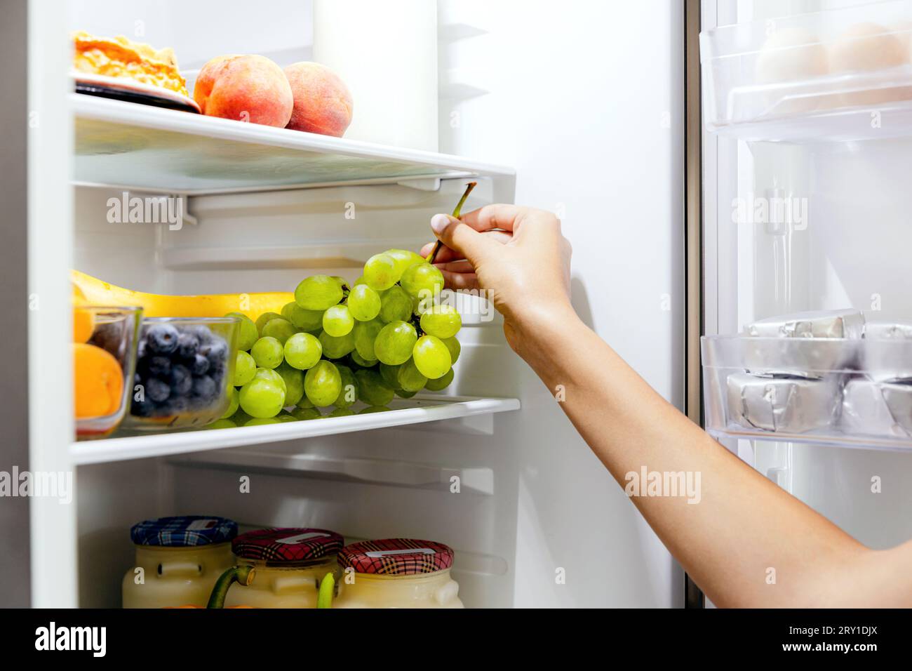 Woman hand taking, grabbing or picks up green bunch of grapes out of open refrigerator shelf or fridge drawer full of fruits, blueberries, bottles wit Stock Photo