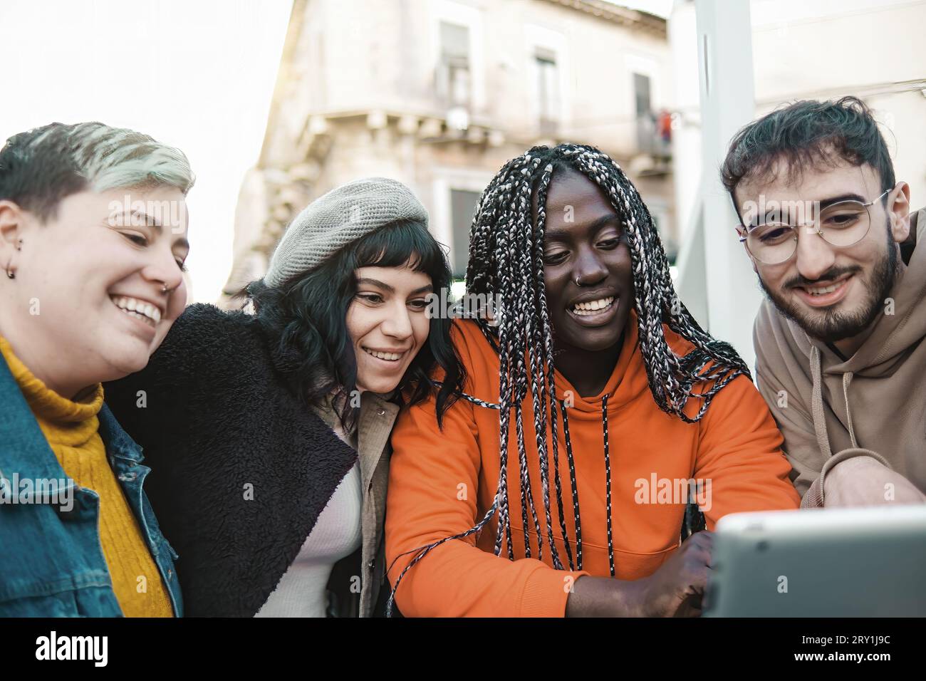 A diverse group of young adults, including a non-binary individual with colorful hair and nose piercings, gathers outdoors, laughing and engaging with Stock Photo