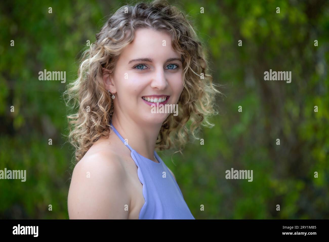 Head and shoulders portrait of a smiling, young Caucasian woman with curly blonde hair and blue eyes, outdoors in Summer Stock Photo