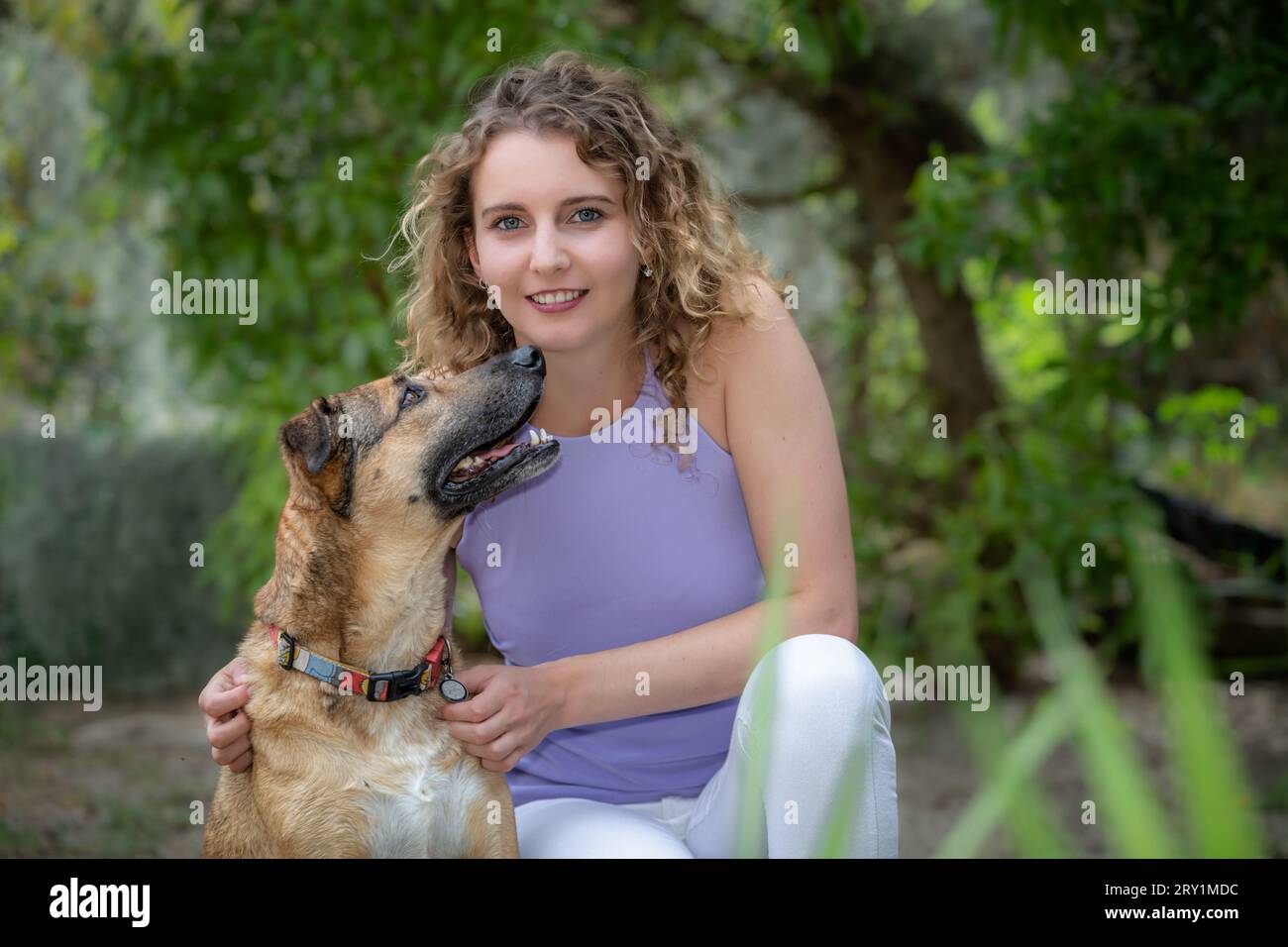 Head and shoulders portrait of a smiling, young Caucasian woman with curly blonde hair and blue eyes, outdoors in Summer with her pet dog Stock Photo