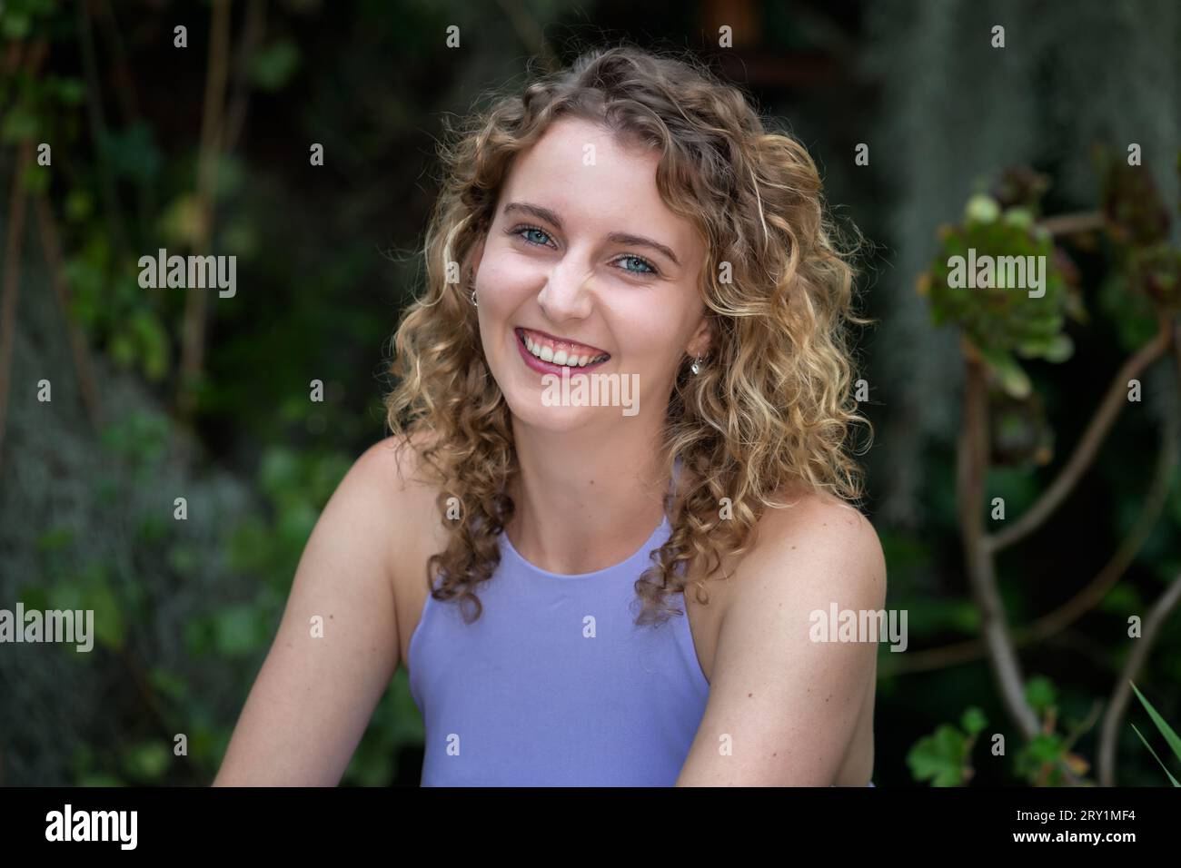 Head and shoulders portrait of a smiling, young Caucasian woman with curly blonde hair and blue eyes, outdoors in Summer Stock Photo