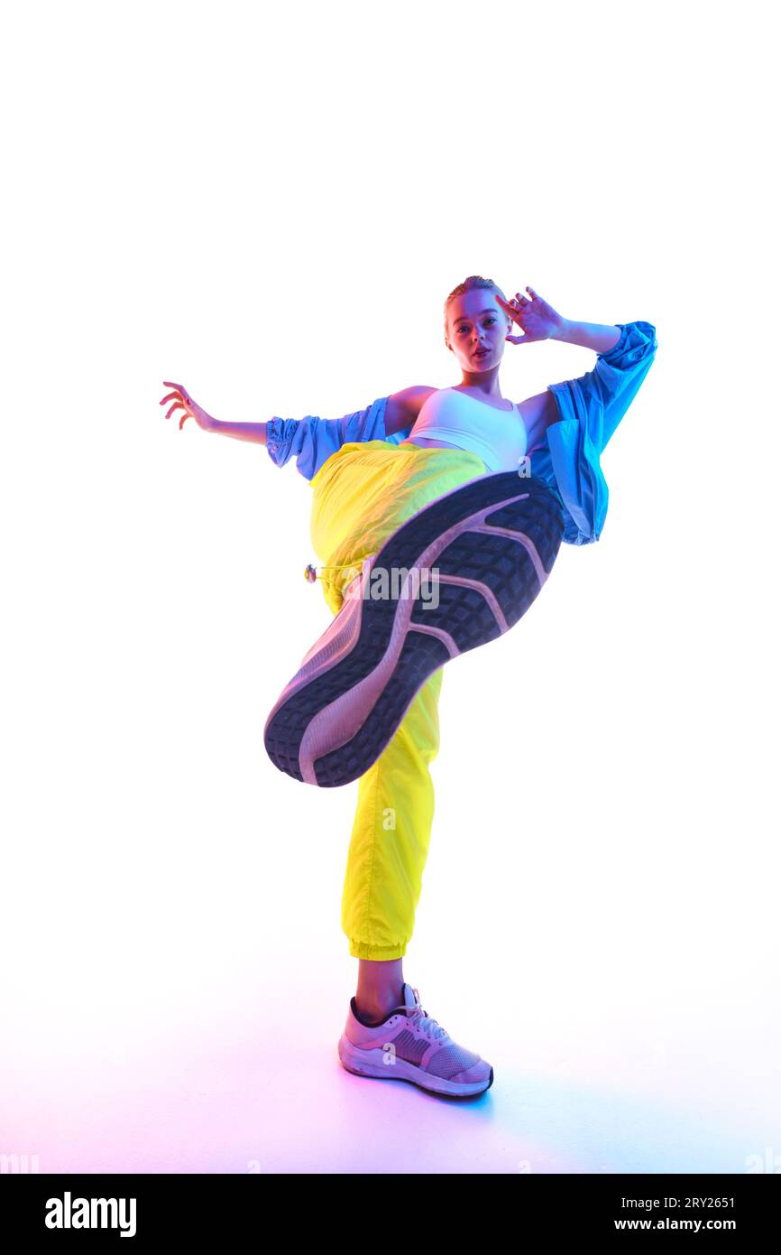 From below full body of attractive young woman training, dancing in trendy sport outfit and sneakers looking at camera in neon light. Stock Photo