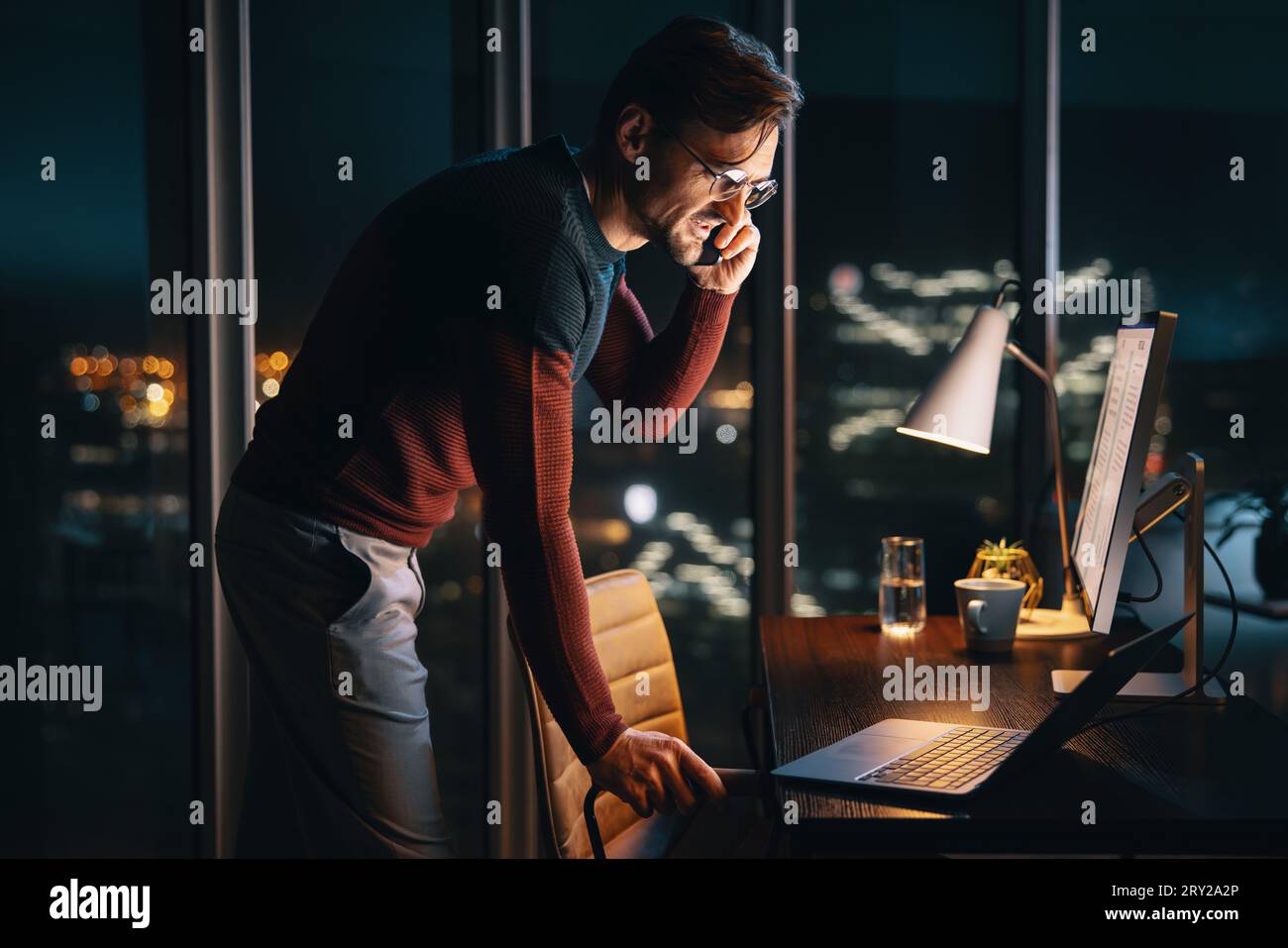 A dedicated business person, working late from a home office, discusses a project on a phone call. This caucasian male entrepreneur utilizes technolog Stock Photo