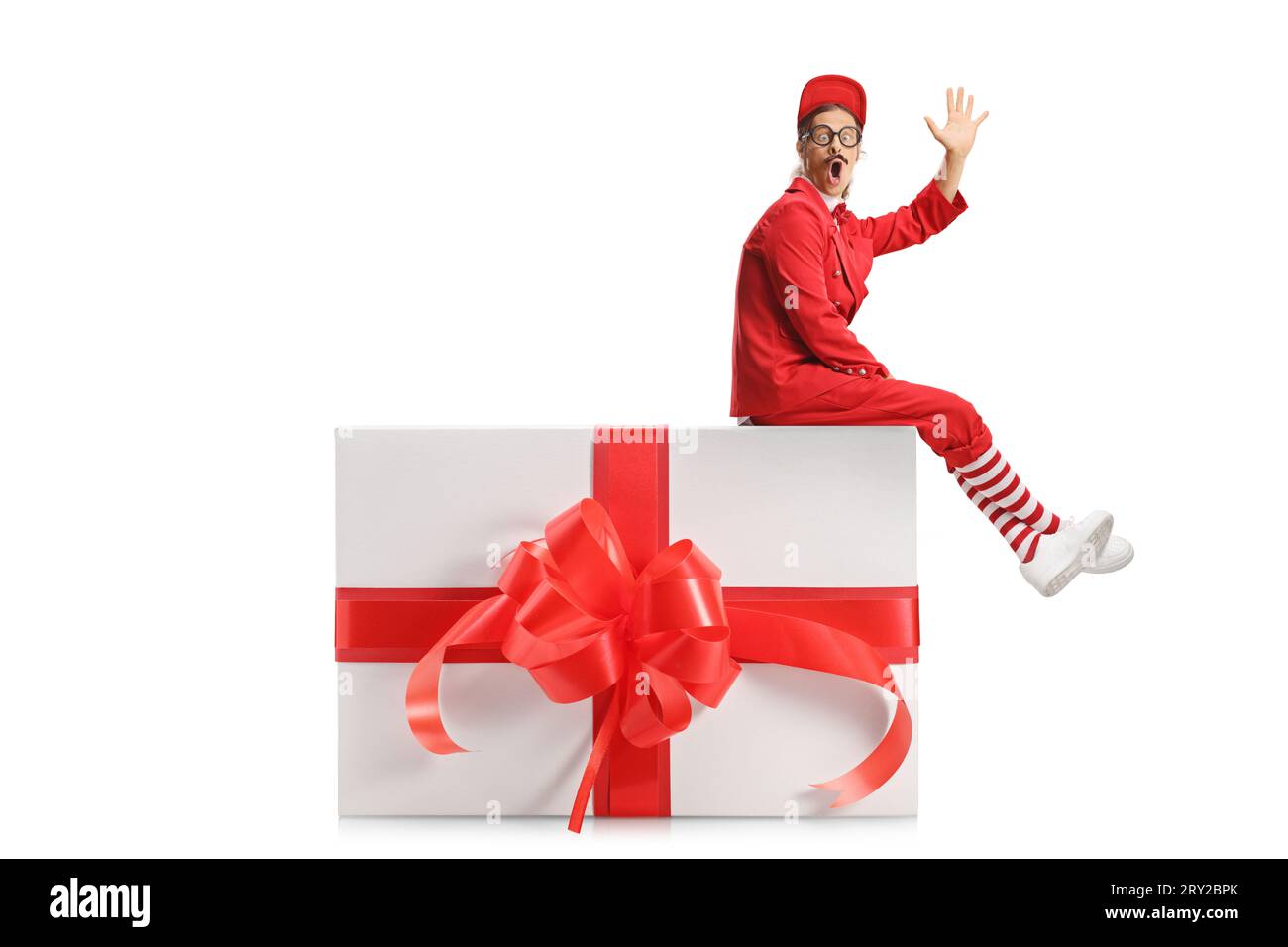 Entertainer in a red suit sitting on a big present box and waving isolated on white background Stock Photo