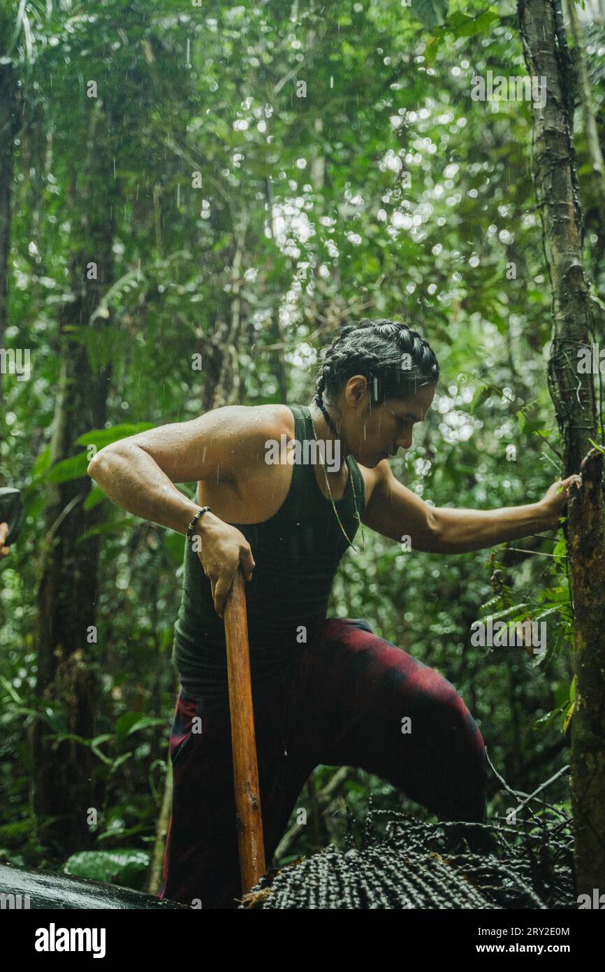 Side view of focused ethnic man with braided hair working with stick while standing in lush greenery of Amazon rainforest Stock Photo