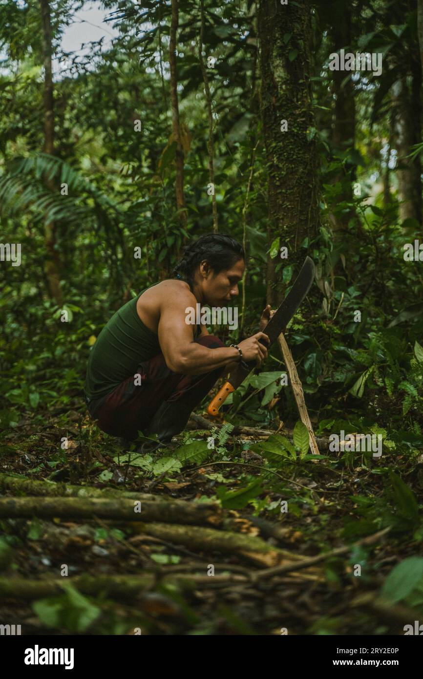 Side view of male using machete knife to cut wood stick working on ground in tropical rainforest Stock Photo