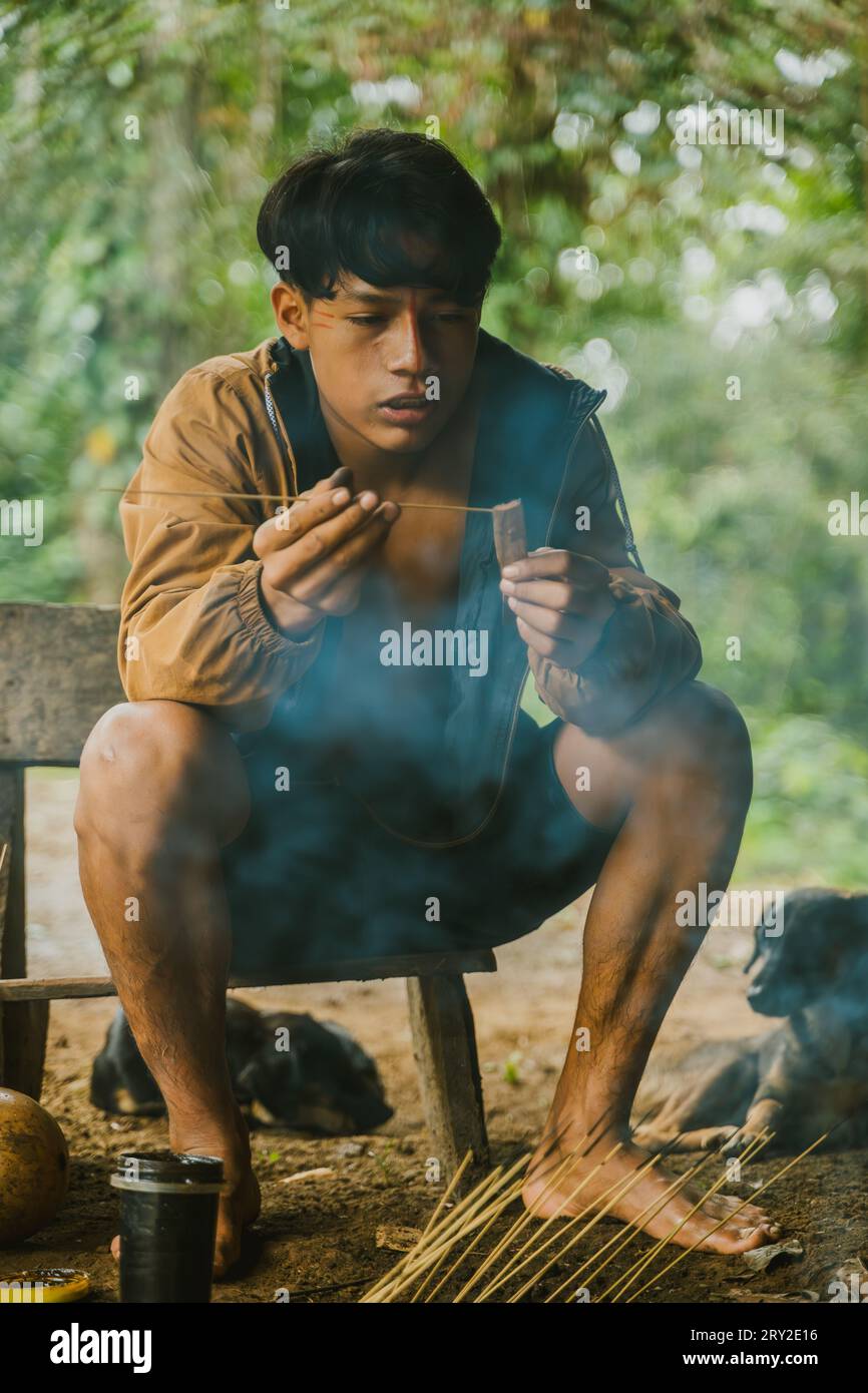 Full body of ethnic young man sitting in smoke and dipping darts in curare poison making traditional weapon for Huaorani tribe Stock Photo