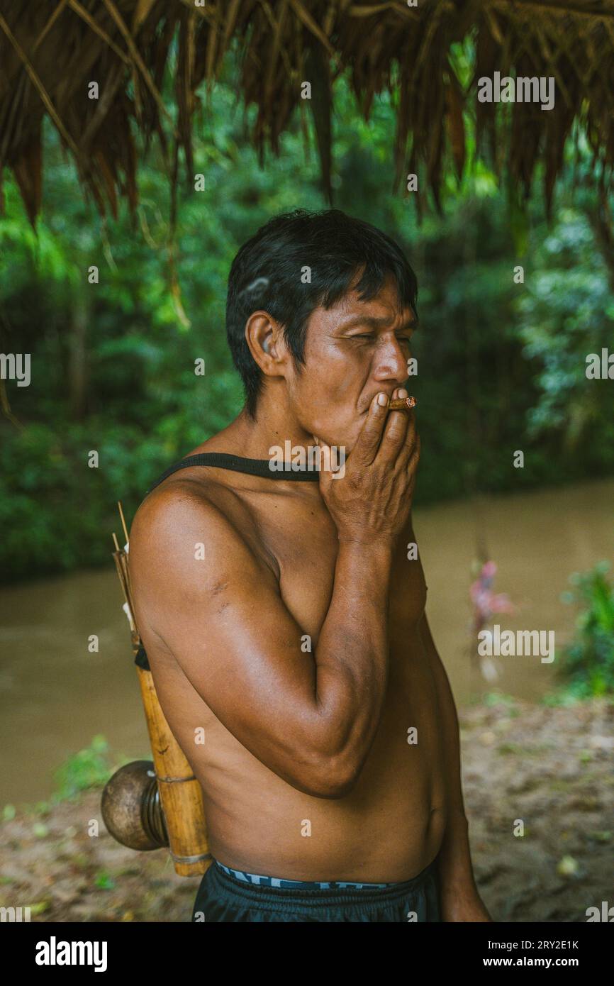 Side view of relaxing adult man of local tribe resting on shore of river in Amazon rainforest and smoking in daylight Stock Photo