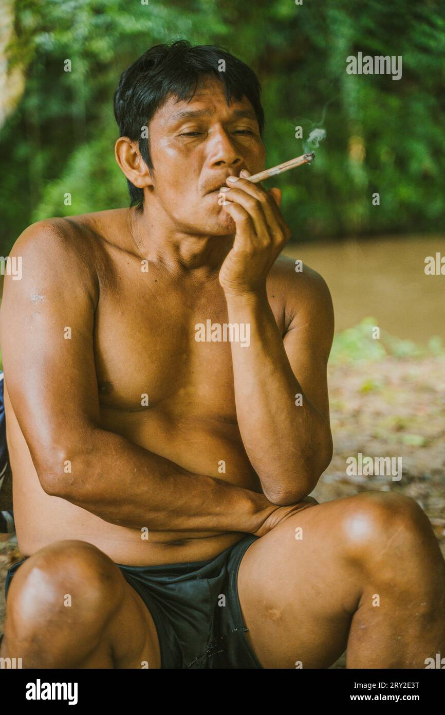Relaxing adult man of local tribe resting sitting on shore of river in Amazon rainforest and smoking in daylight Stock Photo