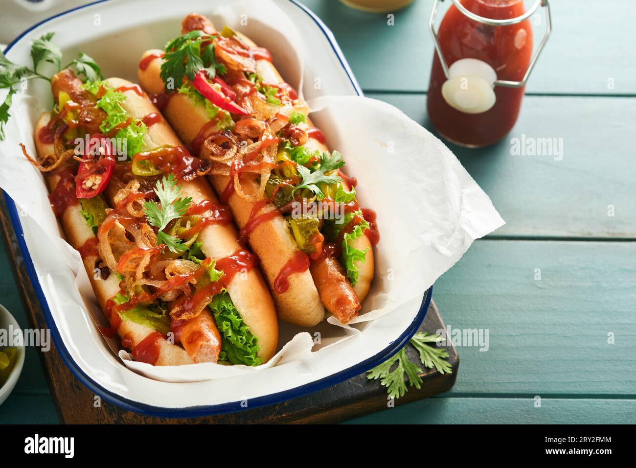 Delicious grilled hotdog with pickled cucumbers, chili peppers, caramelized onions, ketchup, mustard in craft paper on blue old wooden background. Hom Stock Photo