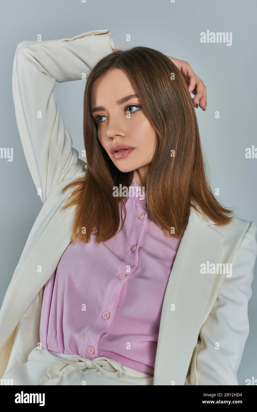 young charming woman in white blazer looking away with hand behind head on grey, fashion and style Stock Photo