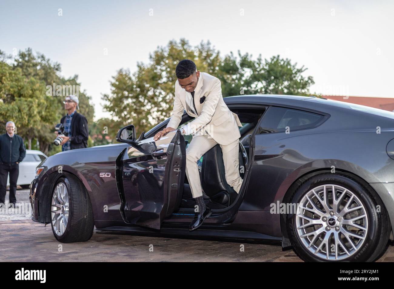 A young mixed-race man, exuding energy and style, emerges energetically from a sleek black sedan at prom Stock Photo