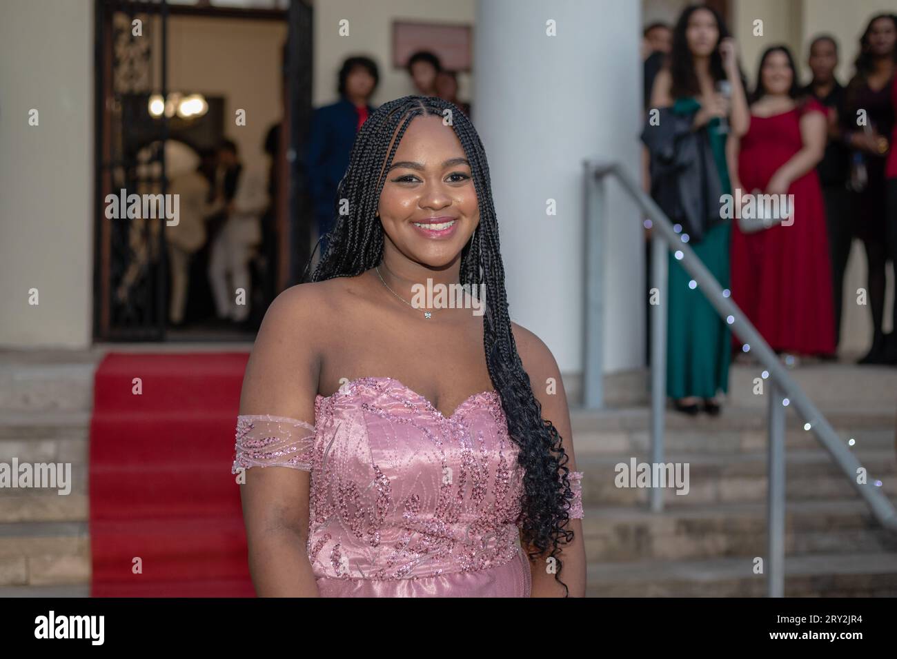 In a close-up shot, a young black woman with braids stands on a red carpeted staircase in a pink cocktail dress, drawing the attention of onlookers Stock Photo