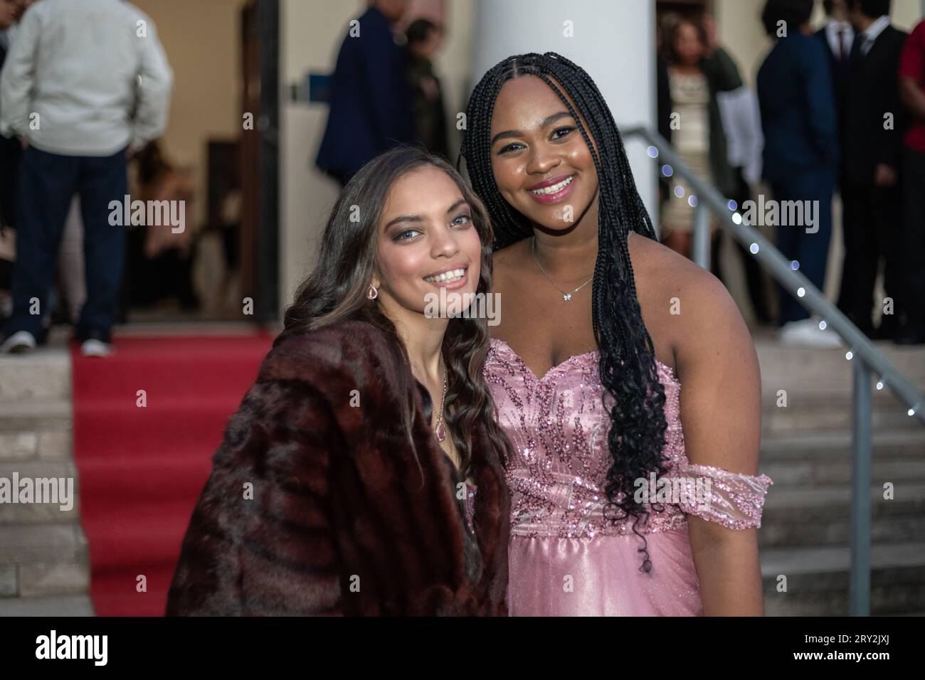 In a close-up shot at the prom, two multiracial friends stand confidently on a red-carpeted staircase, radiating elegance and camaraderie. Stock Photo