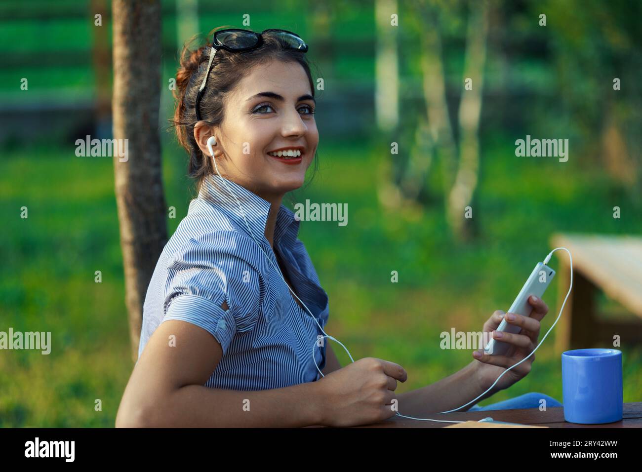 Outdoors, young woman works dynamically, managing phone and laptop. In striped blouse, her hair held by glasses, amidst mountain scenery. Her expressi Stock Photo