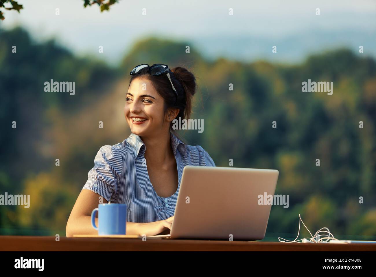 Woman with tied hair, using glasses as a hairband, works outdoors with her laptop, amidst greenery and mountains Stock Photo