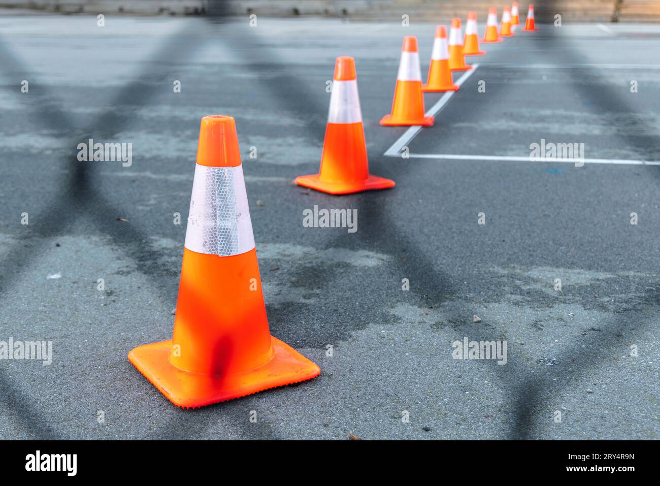 Orange and white traffic cones in a row fading into distance against black tarmac; foreground is a wire fence out of focus. Shot in Vancouver, Canada Stock Photo