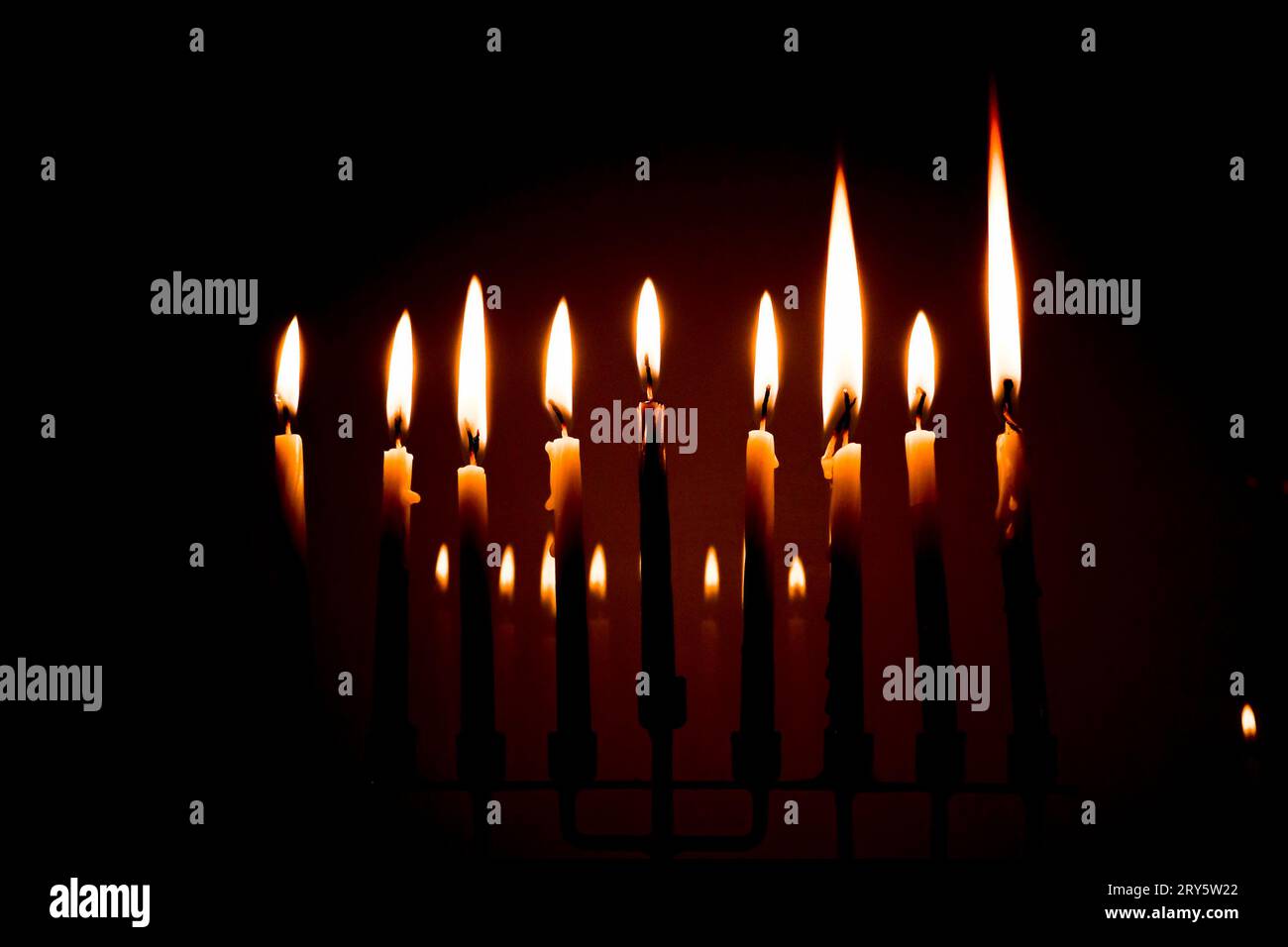 A full menorah on the last night of Hanukkah. Eight white candles and the center is blue. All candles are lit in a darkroom and reflecting in the back Stock Photo