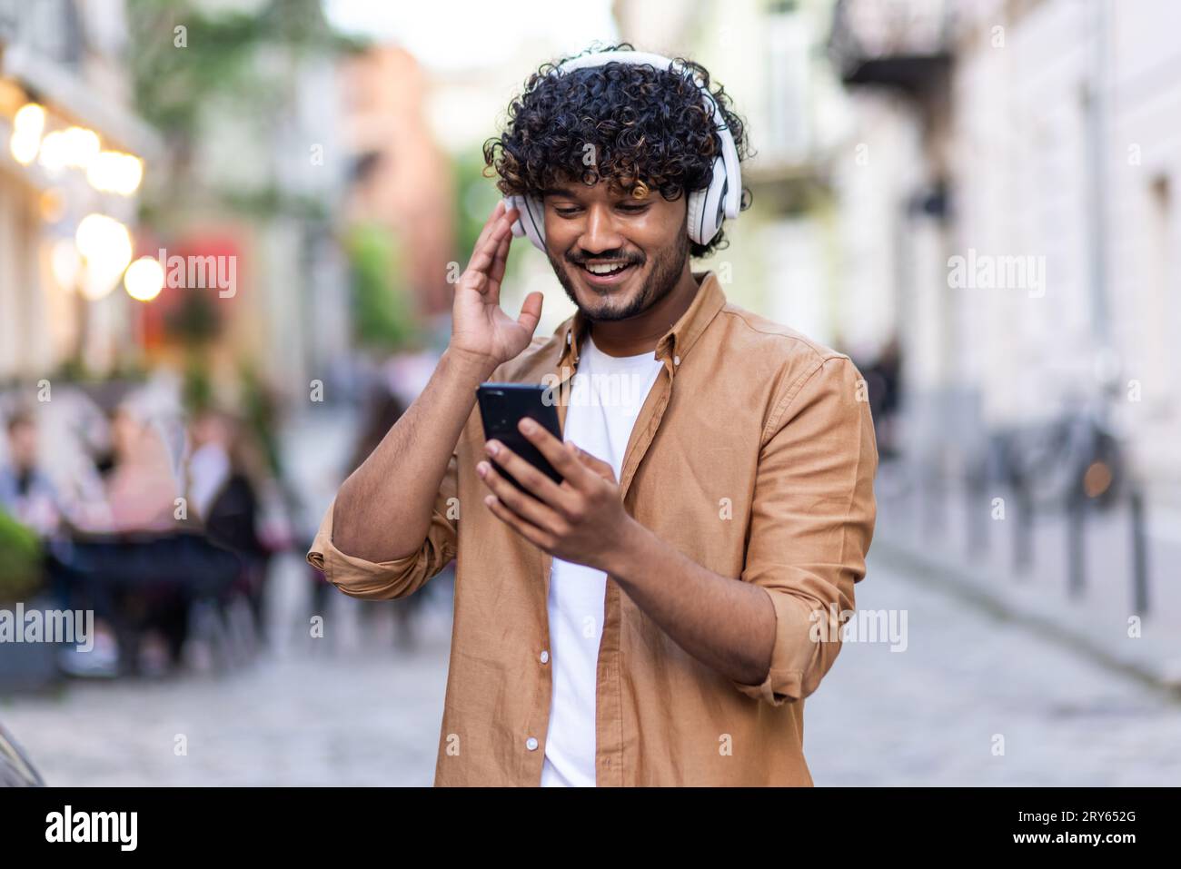 Young Indian man standing on city street wearing headphones and phone and enjoying music, smiling. Stock Photo
