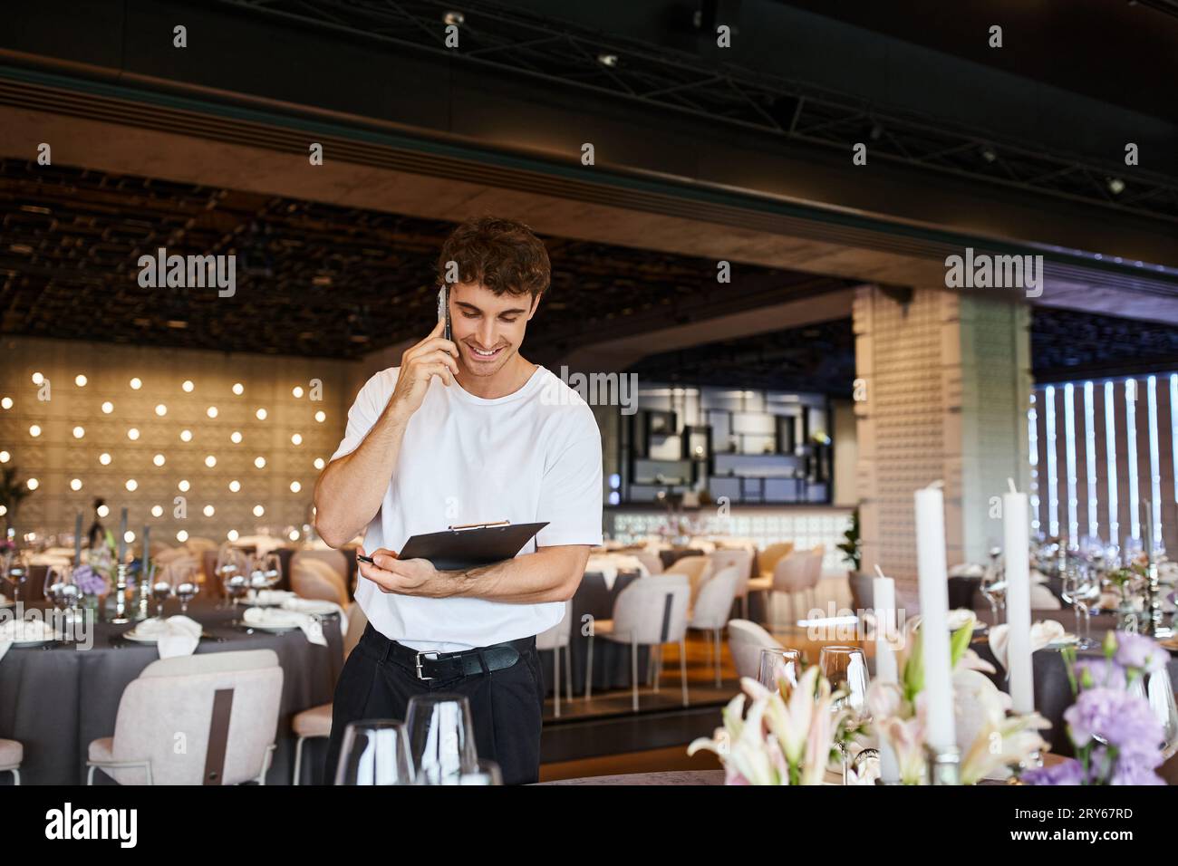 positive event organizer with clipboard talking on mobile phone near festive tables in banquet