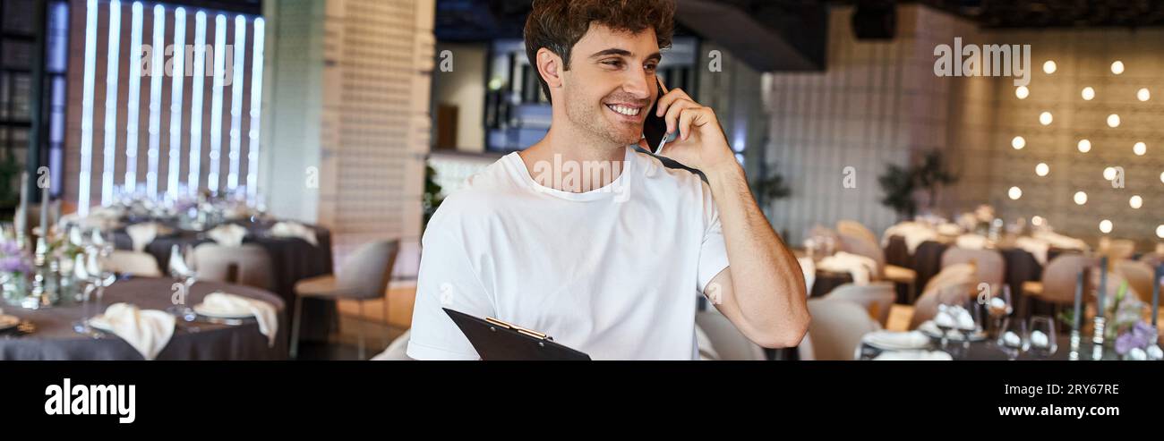 happy event manager with clipboard talking on smartphone near festive tables in banquet hall, banner Stock Photo