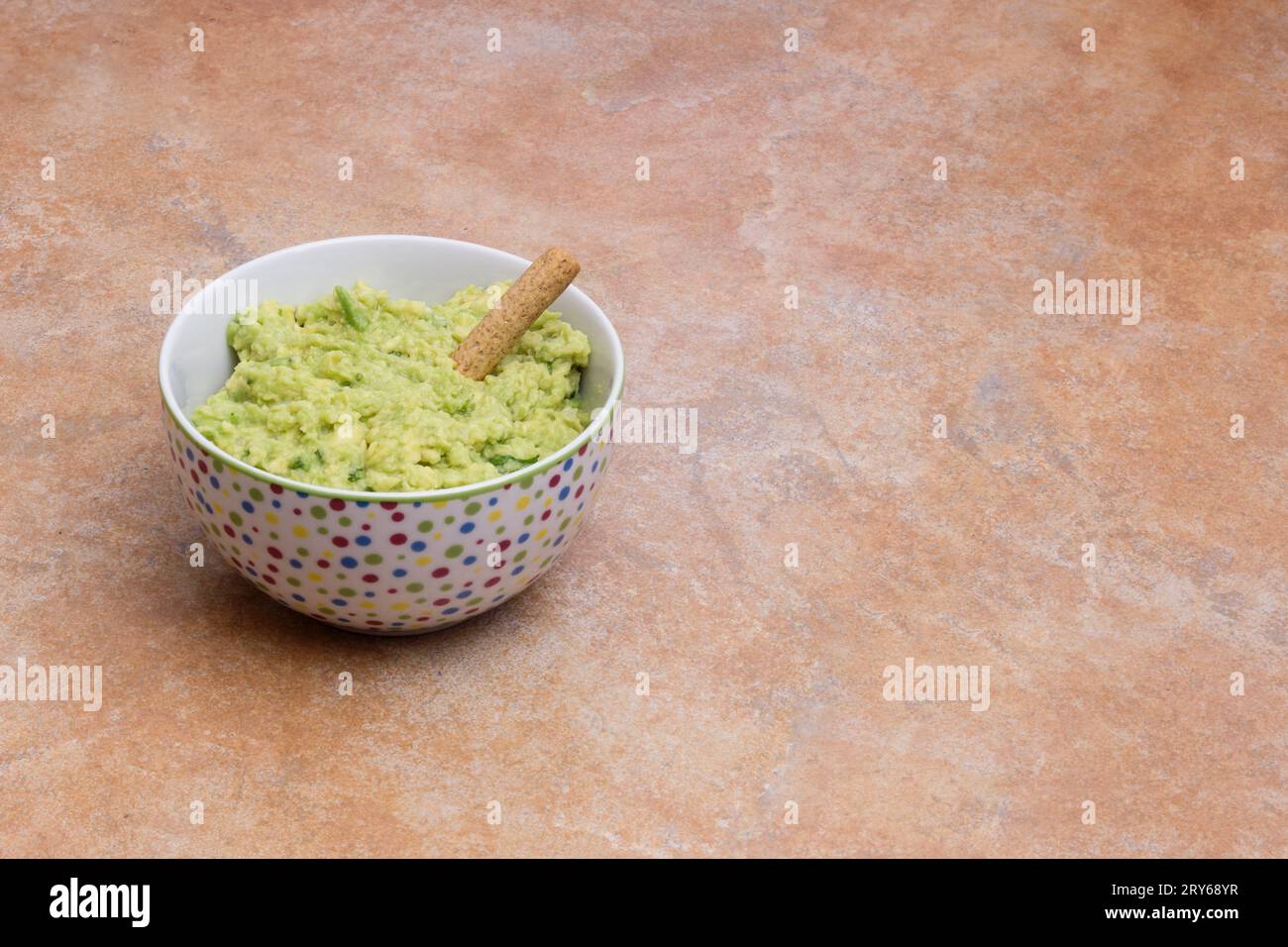 A long shot of a stone-tiled countertop with a bowl of homemade guacamole dip, decorated with colorful dots with copy space. Traditional food. Stock Photo