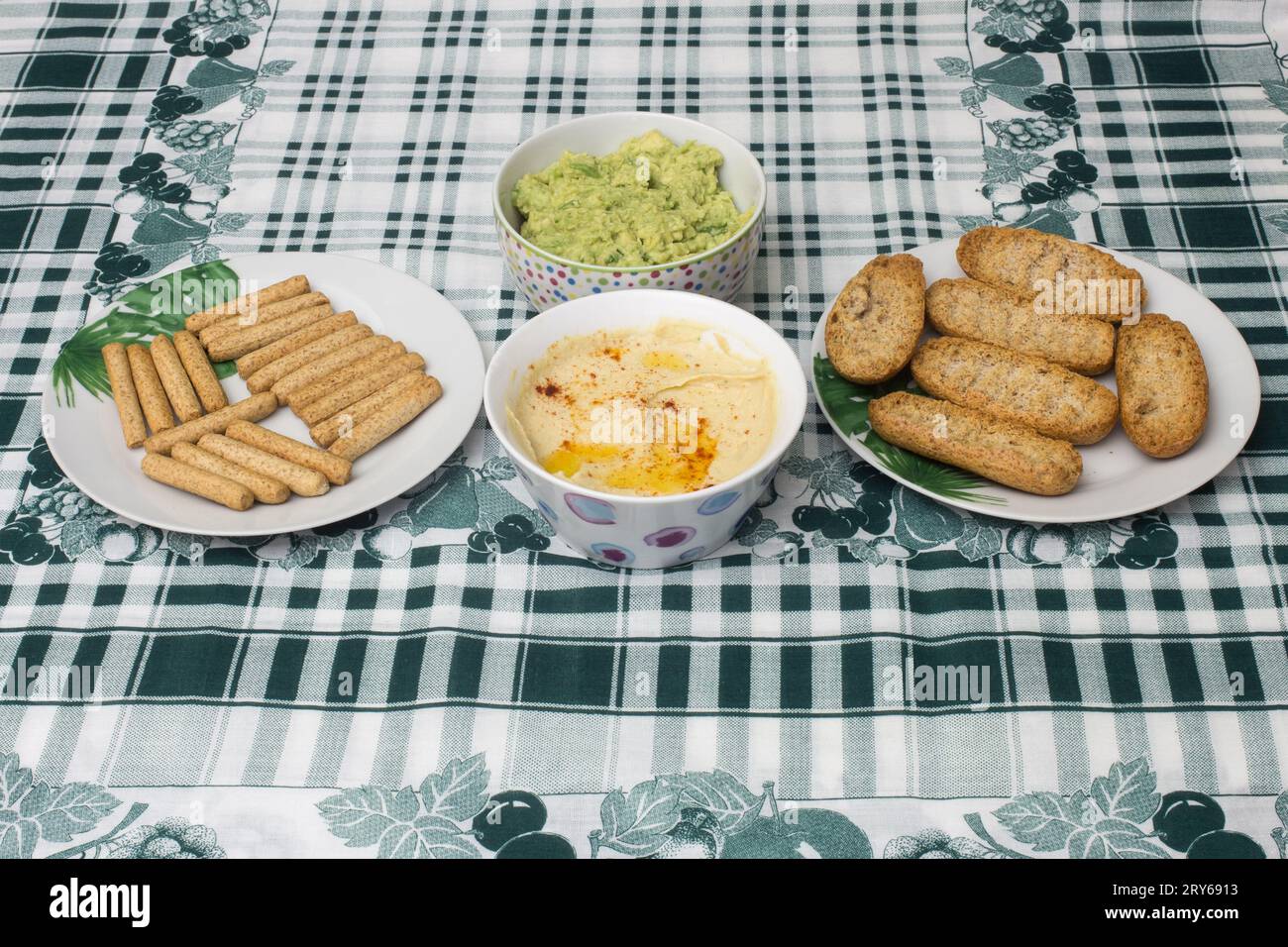 A tablecloth with geometric patterns and elegant leaves decorates the table, where two bowls of hummus and guacamole are served with whole wheat toast Stock Photo
