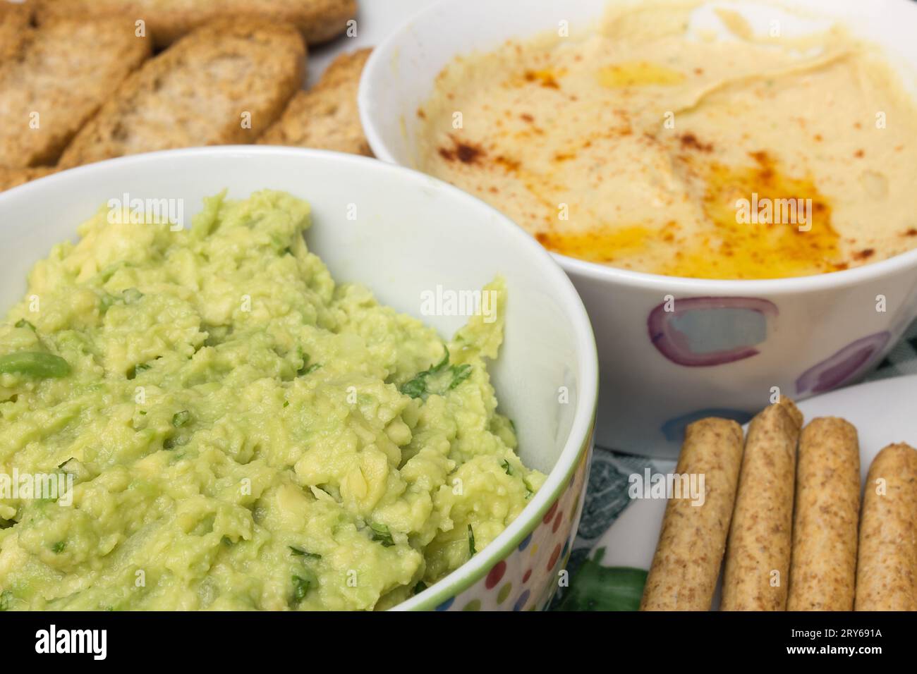 A close-up shot of a bowl of guacamole dip, made with fresh avocado, onion, and cilantro. The background is blurred, but you can see a bowl of hummus, Stock Photo