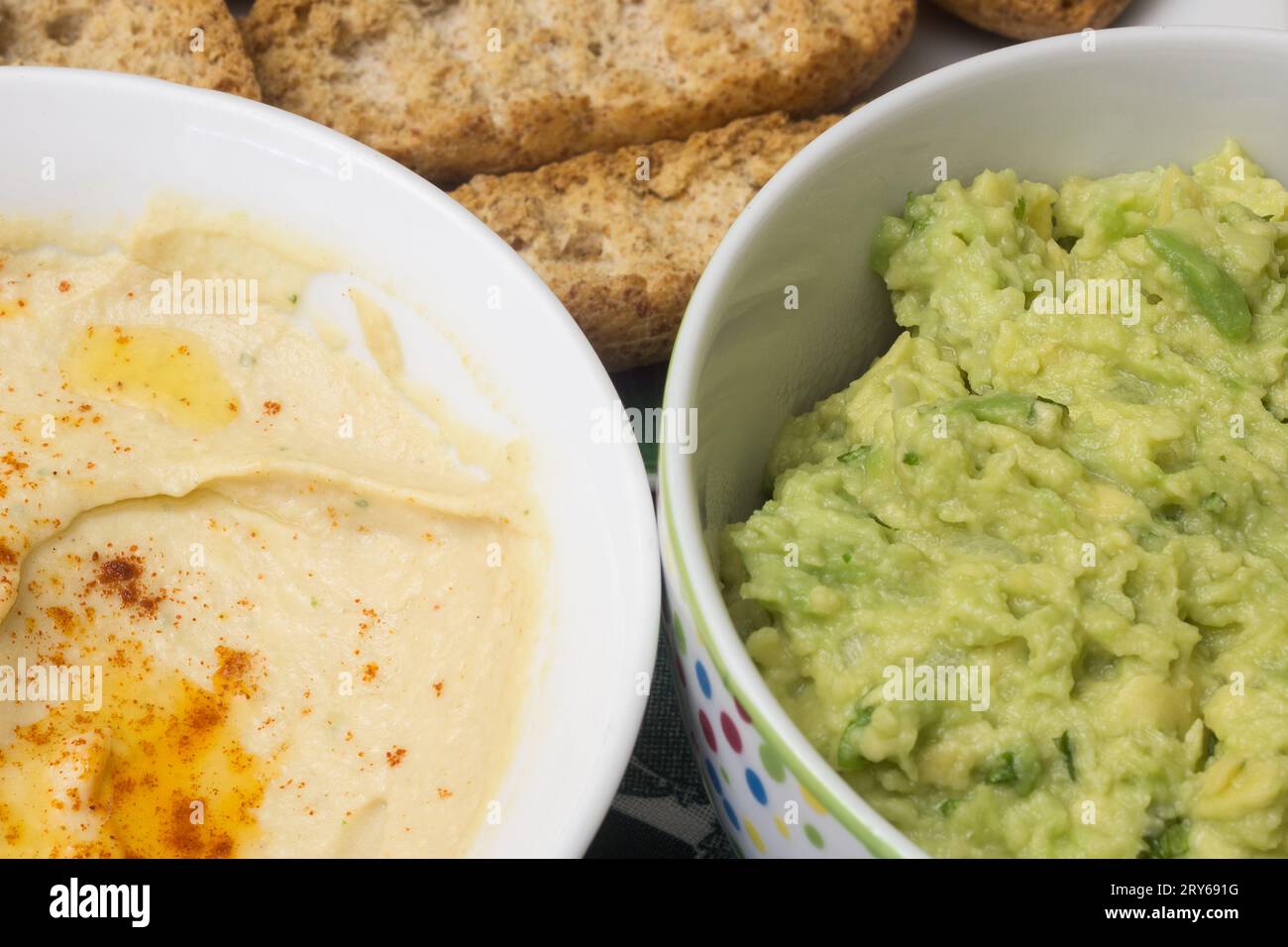 A very close-up shot of two bowls of dip, one with hummus and one with guacamole, both made with fresh ingredients. The breadsticks and toast are out Stock Photo