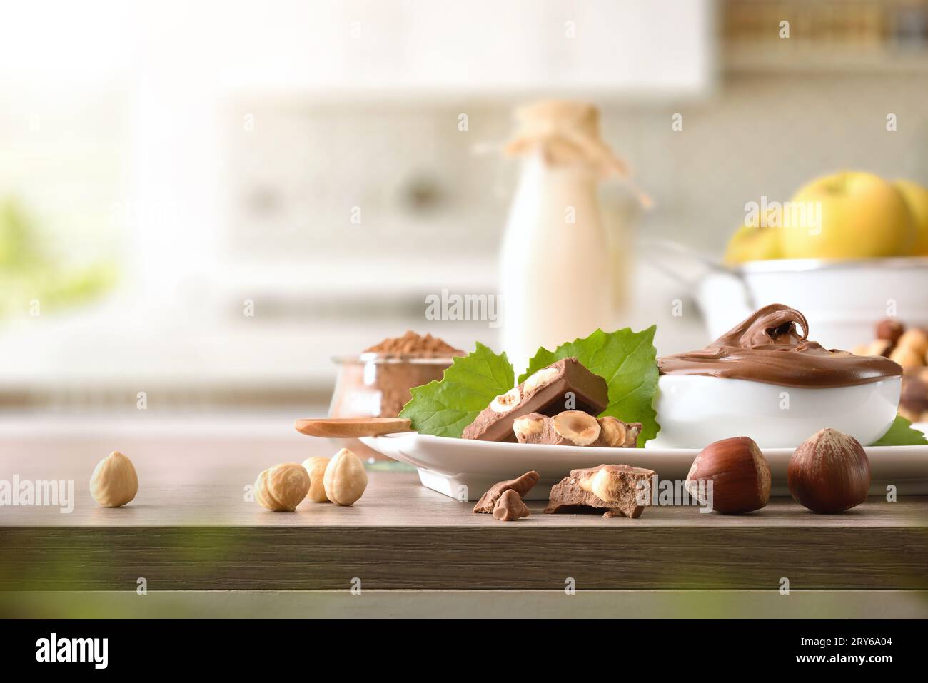Slices and cream of milk chocolate with hazelnuts on wooden table with leaves and nuts around in a kitchen. Front view. Stock Photo