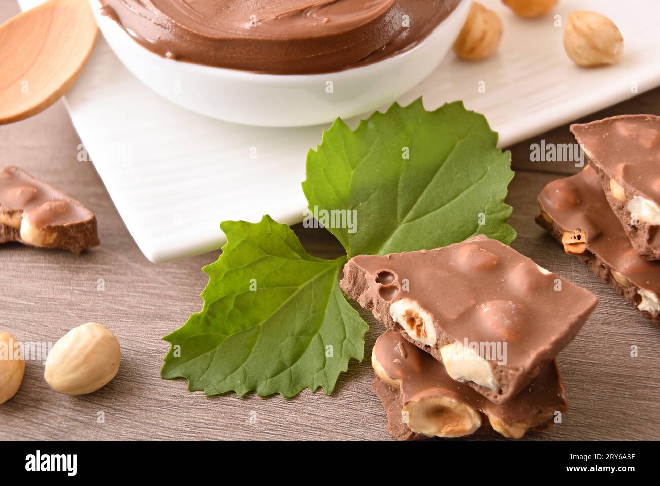 Detail of stack of milk chocolate and hazelnut portions on wooden table with leaves and bowl with praline cream. Elevated view. Stock Photo