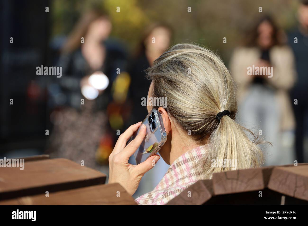 Woman talking on smartphone sitting on a street bench on blurred people background. Using mobile phone in autumn city Stock Photo