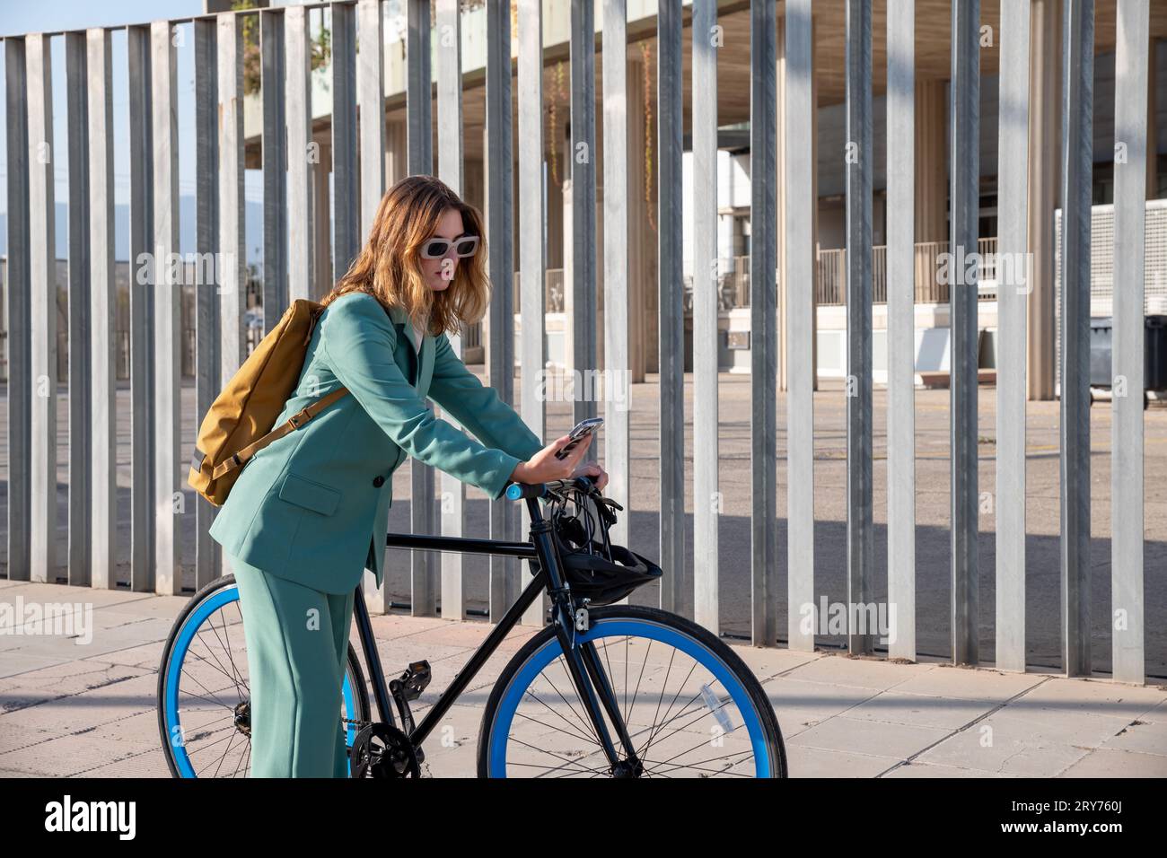 Young empowered ginger-haired woman in green suit using smartphone standing with fixie at the financial district with modern building on the backgroun Stock Photo