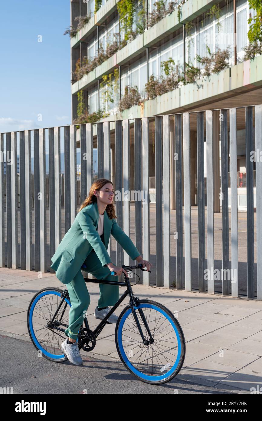 Young empowered ginger-haired woman in green suit riding a fixie at the financial district with modern building on the background Stock Photo