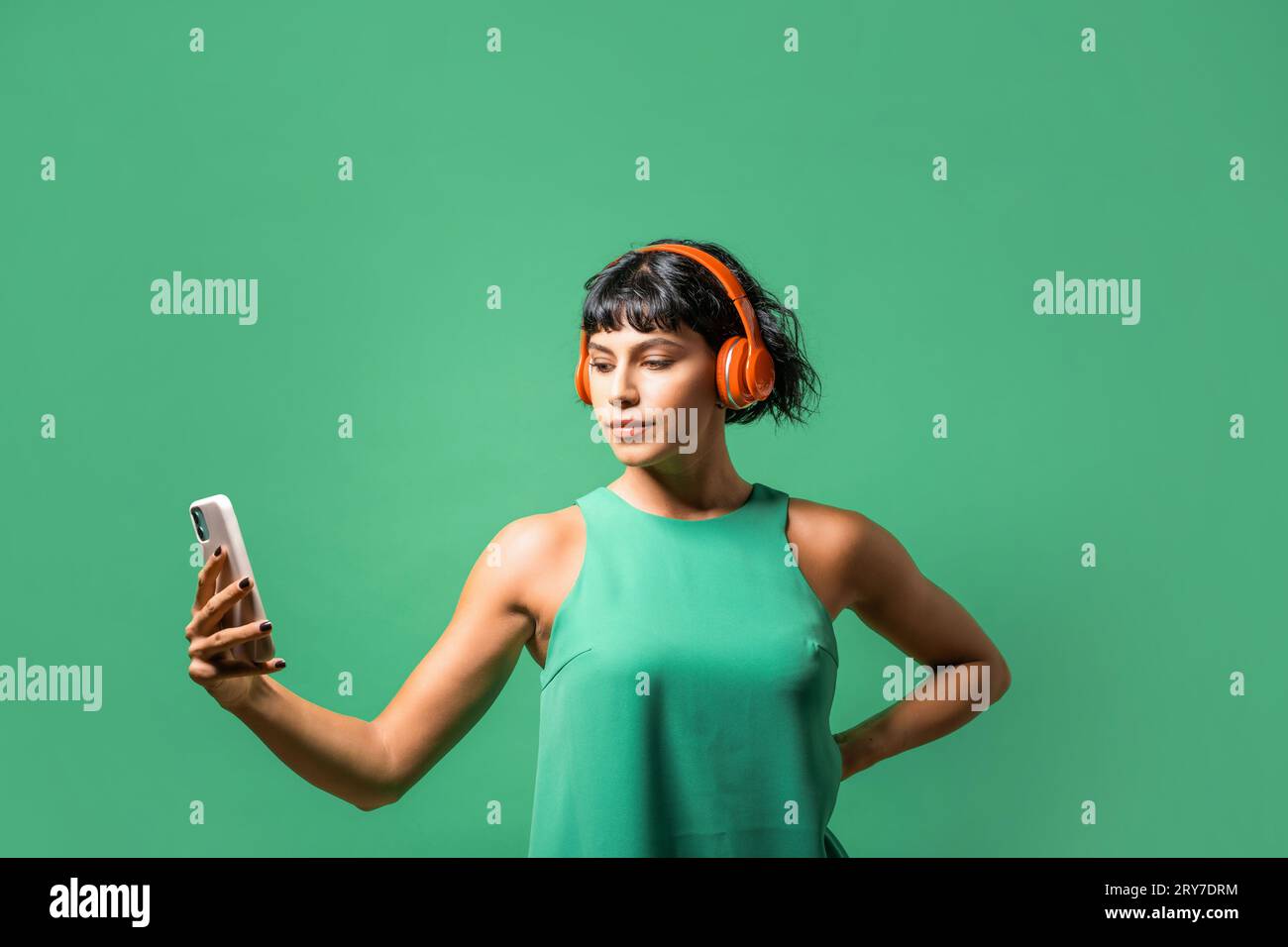 Portrait of a young woman enjoying music on her phone, wearing a red headphone and green tank top against a matching green background. Stock Photo