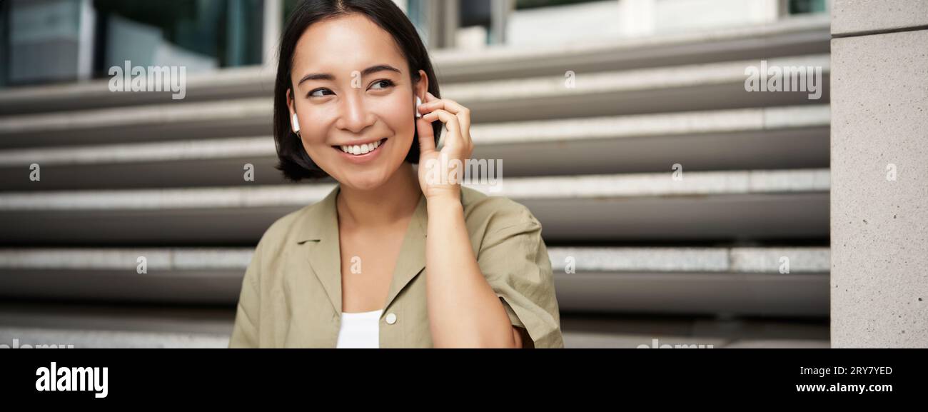 Close up portrait, street photo of asian woman listening music in wireless headphones, smiling, sitting on stairs Stock Photo