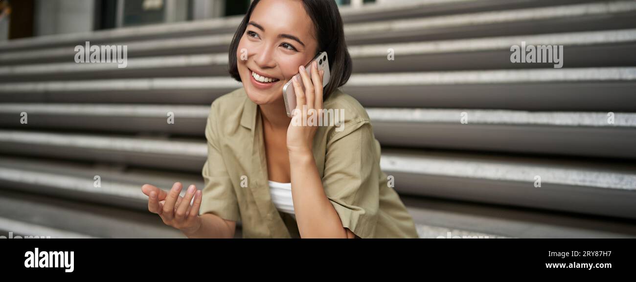 Close up portrait of smiling asian girl talks on mobile phone, sits outside on street stairs. Young woman calling friend on smartphone Stock Photo