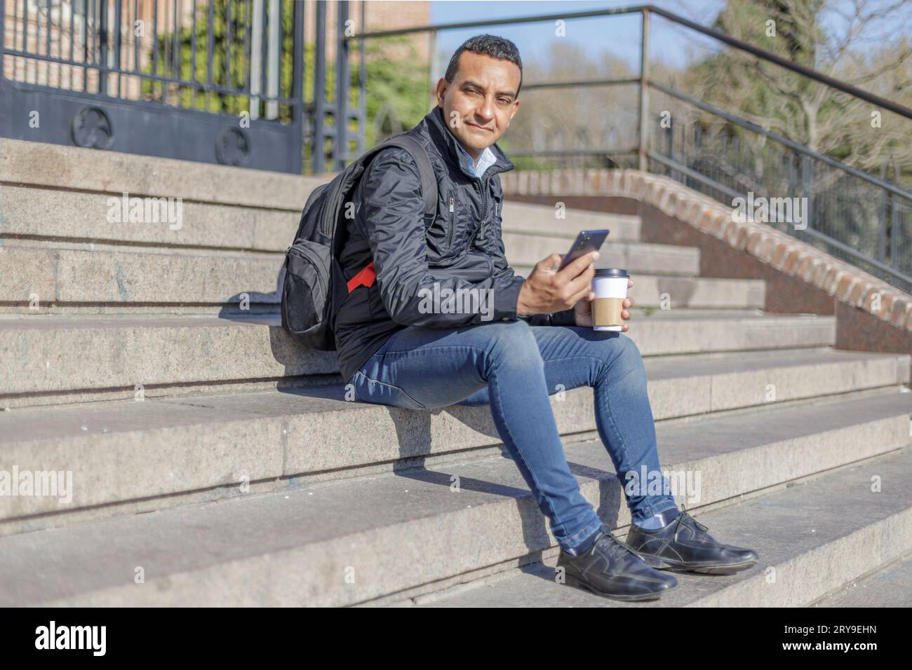 Latino man sitting on some stairs with a mobile phone and a disposable cup of coffee. Stock Photo