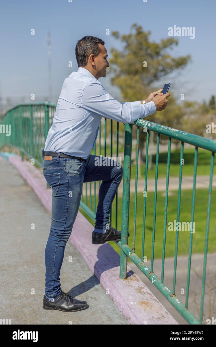 Latin man using his mobile phone leaning on the railing of a pedestrian bridge. Stock Photo