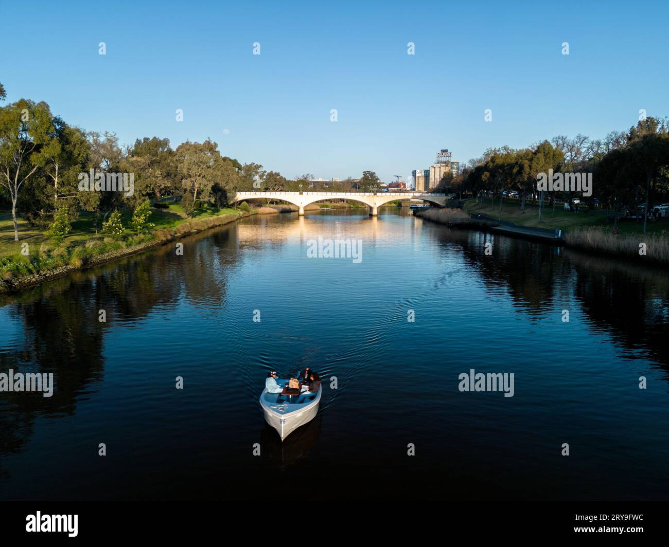A view of a small boat motoring along the Yarra River in Melbourne, taken in the late afternoon with trees framing the river and a large bridge in the Stock Photo