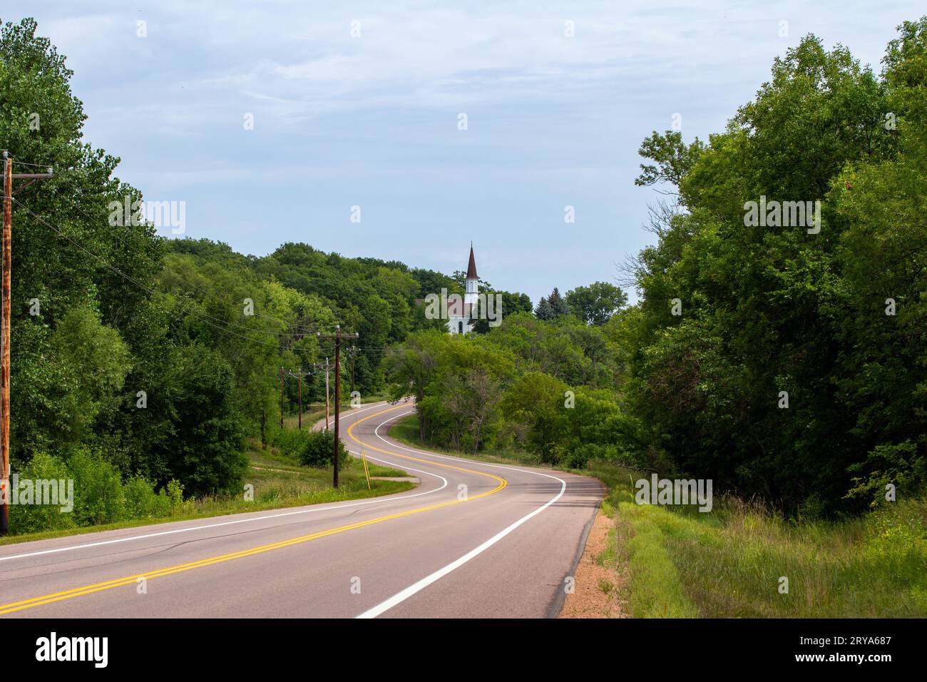 Landscape view of a picturesque white wooden country church on a rural hillside, with view of a winding highway Stock Photo