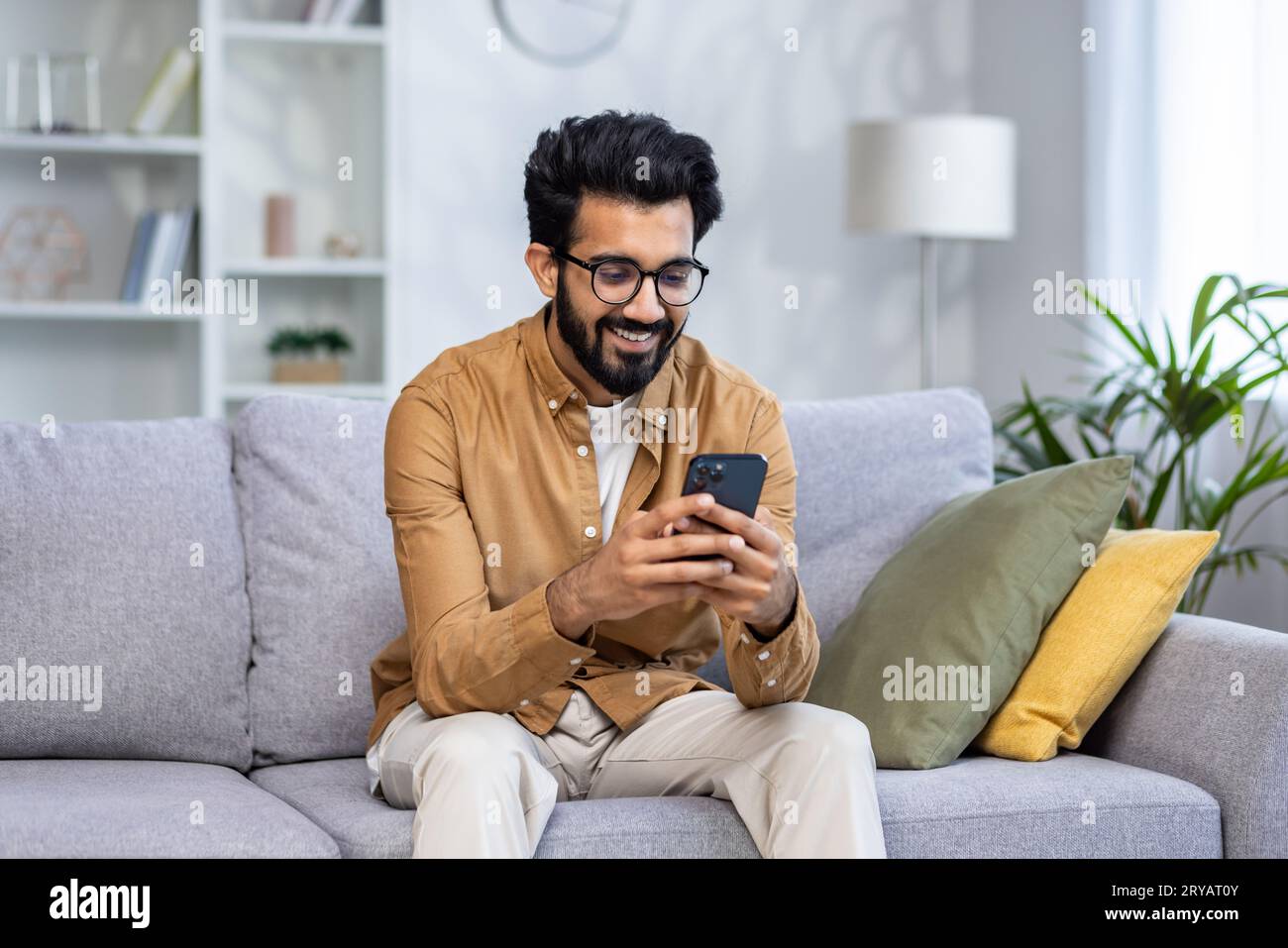 Young hispanic man sitting at home on sofa in living room of house, happy and smiling man holding phone, using smartphone app, typing message and watching video, online shopping. Stock Photo