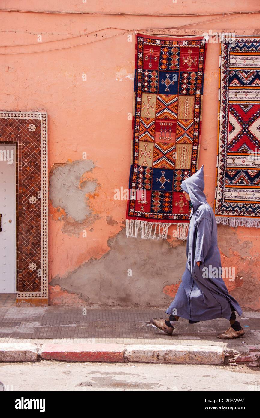 local man in Djellaba walks past hanging rugs in the streets of Marrakech Morocco March 2012 Stock Photo