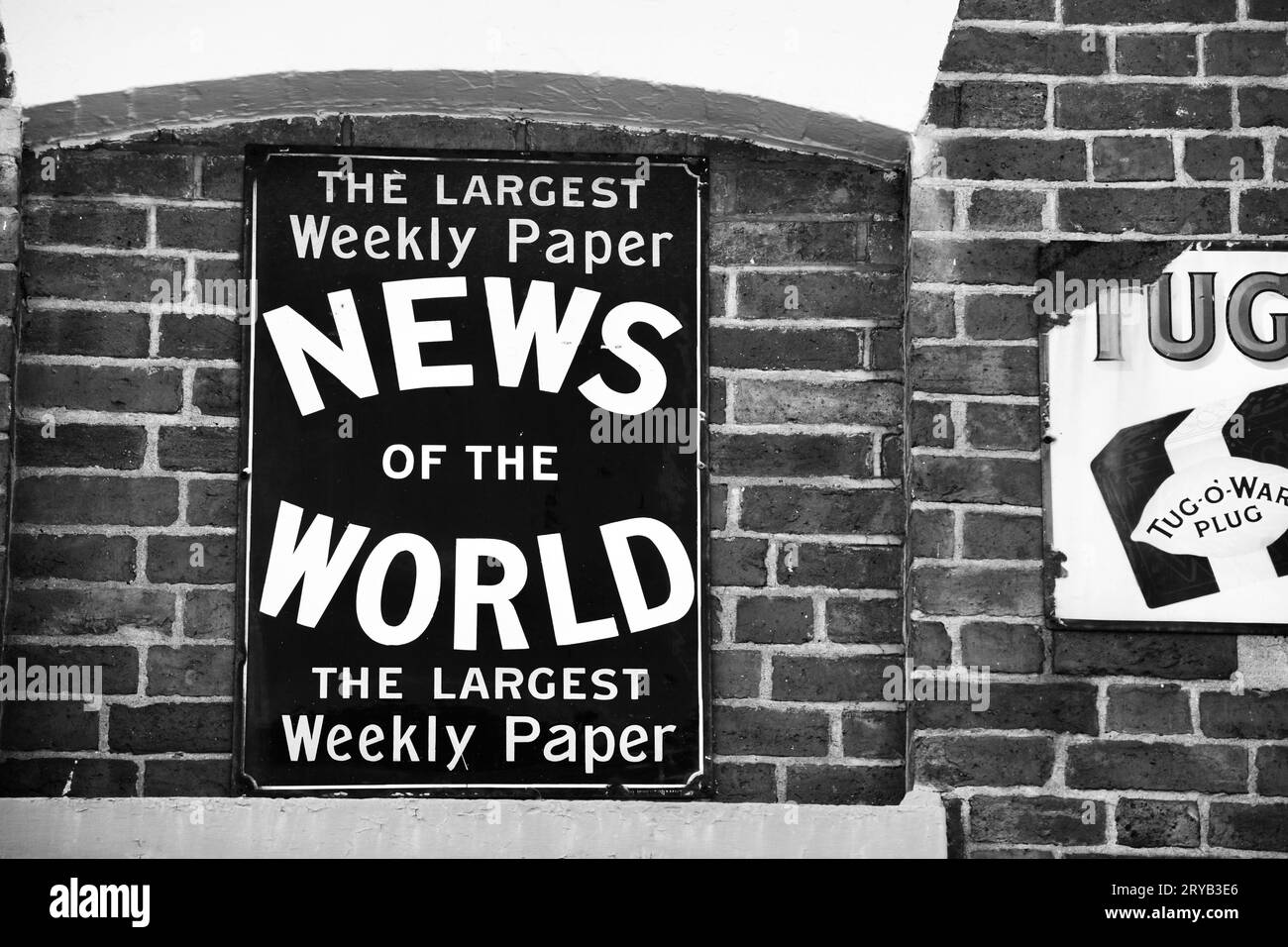 Black and white photo of a vintage, metal advertising sign displayed on a brick wall advertising the News of the World newspaper. Stock Photo