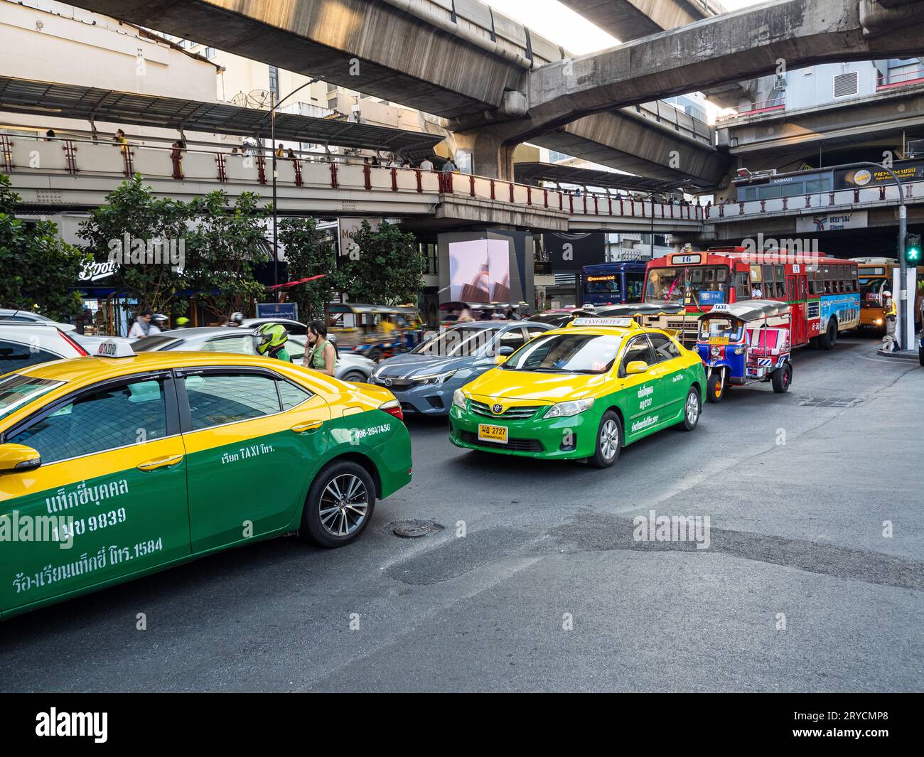 A lively Bangkok street, bustling with green and yellow taxis, buses, cars, and a tuk-tuk beneath the Skytrain's concrete supports. Stock Photo